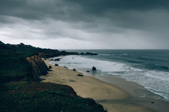 Capturing the atmosphere of a stormy day by the ocean, this image highlights the rugged cliffs, sandy beach, and turbulent waves under an overcast sky. Suitable for background images in travel websites, blogs, or brochures focusing on nature, coastal tourism, or weather-related content.