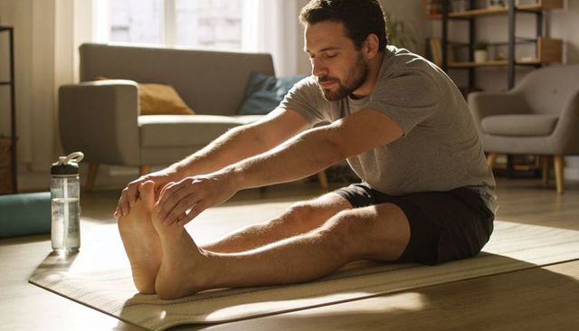 Young man stretching on yoga mat in sunlit living room reaching toes for hamstring stretch