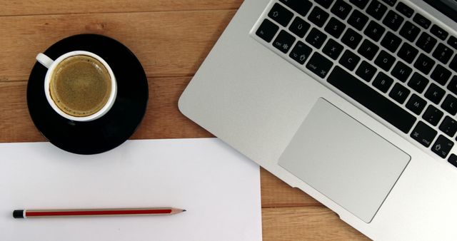 A cup of coffee sits next to a laptop and a blank sheet of paper with a pencil on a wooden desk, with copy space. It suggests a setting poised for productivity, perhaps in a home office or workspace.