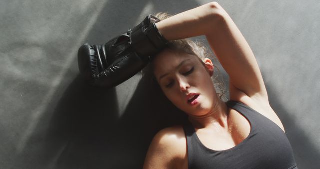 Woman in black boxing gloves and athletic wear resting with one arm on her forehead, showing signs of exhaustion after a rigorous training session. This is suitable for promoting fitness programs, sports training articles, boxing equipment, motivation campaigns, or healthy lifestyle blogs.