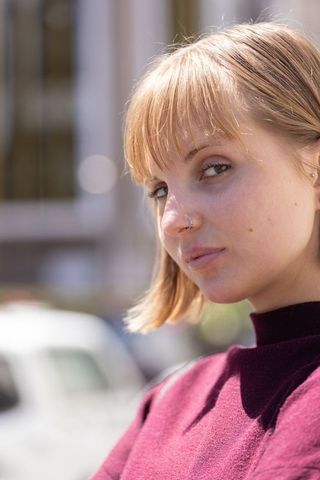 Confident young woman with nose hoop and maroon sweater posing in sunlit urban portrait