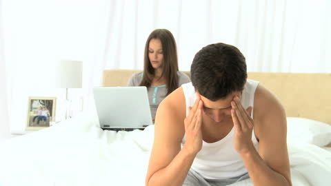 Man Sitting on Bed with Woman in Background Using Laptop