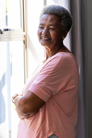Senior african woman gazing out window smiling in pink top with soft natural sunlight