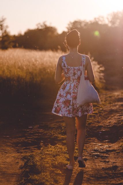 Young woman is walking alone down a rural path at sundown, wearing a floral summer dress and carrying a bag. Ideal for concepts of tranquility, solitude, nature walks, or summer activities advertisement.
