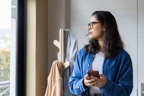 Asian woman standing by window checking phone wearing denim shirt and tortoiseshell glasses