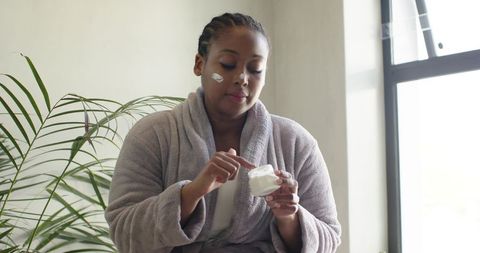 African american woman applying moisturizer in plush robe during morning self-care routine