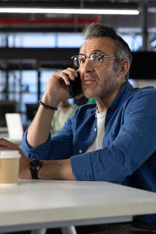 Middle-aged man talking on phone at coworking desk wearing round glasses and wristwatch