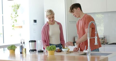 Young couple preparing fresh meal on sunlit wooden kitchen island with blender and herbs