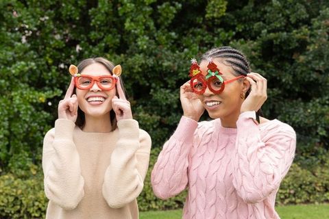 Two diverse women wearing festive novelty glasses laughing in cozy sweaters outdoors