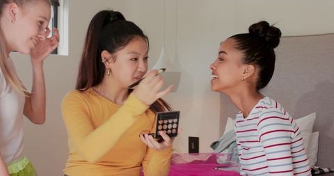 Multicultural friends applying makeup on bed laughing and sharing beauty tips during sleepover