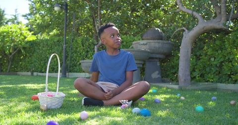 Boy sitting on lawn during colorful easter egg hunt with basket and pastel eggs in sunny backyard