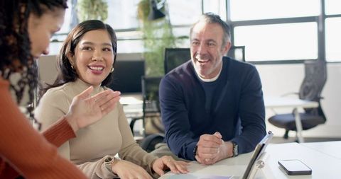 Diverse team collaborating and smiling around laptop in bright modern office with greenery