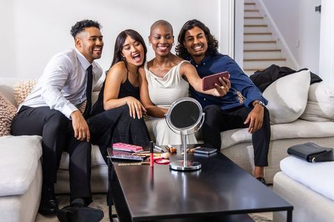 Diverse friends taking joyful selfie on sofa wearing elegant outfits and makeup