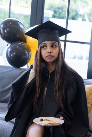 Young graduate wearing mortarboard and gown holding snack plate at graduation reception