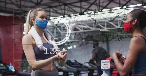 Women wearing masks conversing at gym sanitizer station during cardio session with futuristic hud