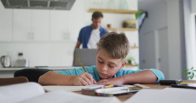 Young boy deeply focused on completing homework at a desk, with a home kitchen backdrop including his father in the background. Ideal for use in articles or advertisements about family life, education, remote learning, parental support in education, and home environments promoting learning.