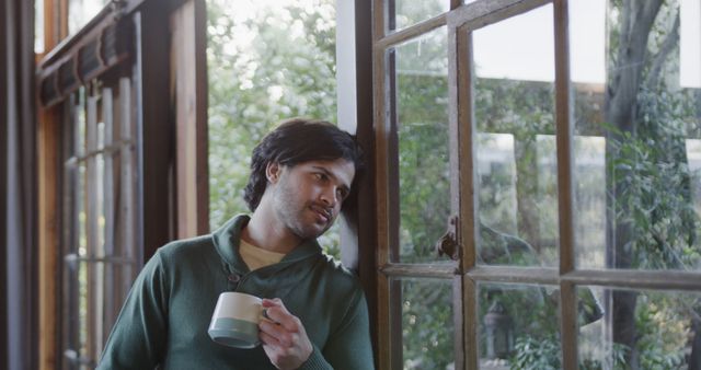 Man holding coffee cup while looking out of window, reflecting and thinking deeply, greenery visible outside. Suitable for themes of contemplation, relaxation, morning routines, peaceful moments, or lifestyle blogs.
