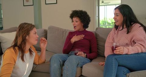 Diverse women laughing while telling stories on sofa holding red drinks in cozy living room