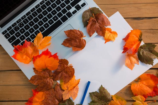 Autumn leaves scattered around paper and laptop on wooden table. Ideal for themes related to fall season, workspaces, creativity, remote work, and nature-inspired office settings.