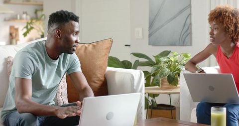 Couple working from home on laptops discussing plans in bright living room with plants