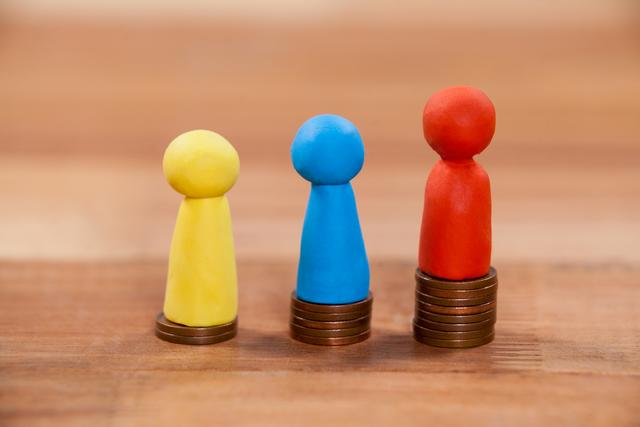Three colorful figurines standing on stacks of coins, representing financial concepts such as wealth distribution, economic disparity, and investment. Useful for illustrating articles on finance, economy, wealth management, and social inequality.