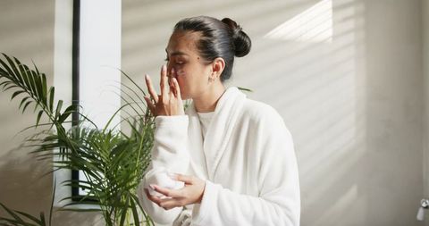 Mid-adult asian woman applying moisturizer in white robe morning skincare natural light