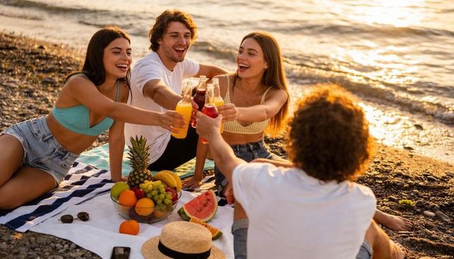 Friends clinking bottles during sunset beach picnic with fresh fruit and relaxed vibes