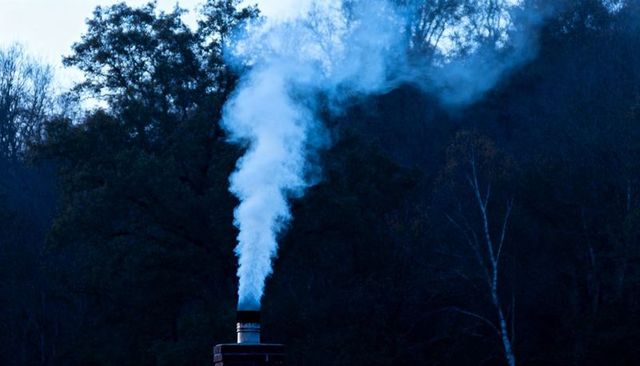 White-blue smoke rising from metal stovepipe on brick chimney against dark birch forest