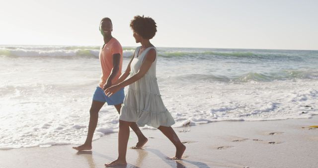 Smiling african american couple holding hands and walking on sunny beach - Download Free Stock Photos Pikwizard.com