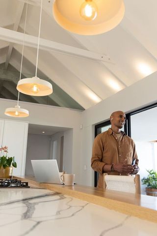 African american man standing in modern kitchen using smartphone, laptop on marble island