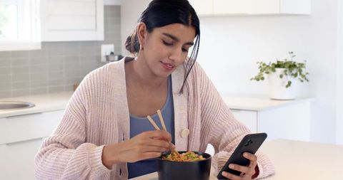 Woman eating noodles while using smartphone in modern kitchen