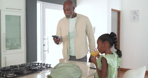 Senior father checking phone while daughter drinks orange juice in modern kitchen