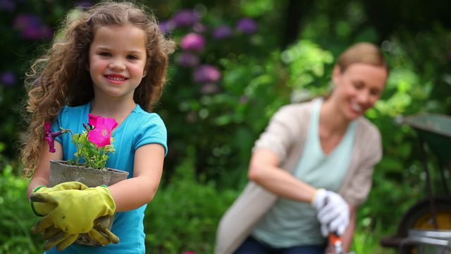 Young girl holding a plant while smiling, with her mother in the background digging in a garden. Perfect for concepts such as family bonding, gardening, outdoor activities, nature, and teamwork.