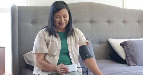 Asian woman checking blood pressure on bed with portable monitor for home wellness routine
