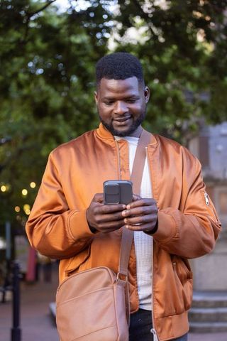 African american man checking smartphone on plaza in rust bomber jacket with crossbody bag