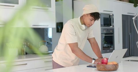 Young man leaning on kitchen island working remotely on laptop with smartphone and fruit bowl