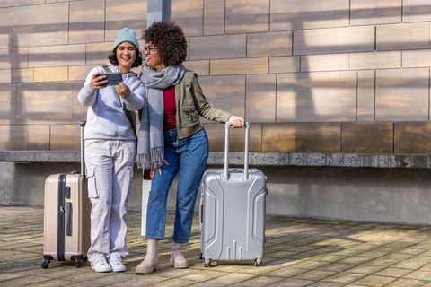 Diverse female friends checking smartphone with suitcases on sunny urban plaza before trip