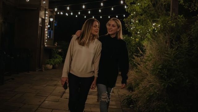 Friends walking arm in arm on lit stone patio at night under string lights cozy sweaters denim