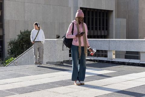 Standing and checking phone on urban plaza wearing pink hoodie, backpack, long braids
