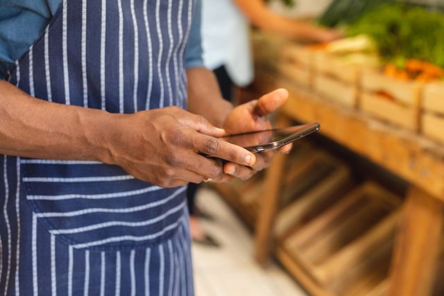 Midsection of african american mid adult male vendor wearing apron using smartphone in store. unaltered, wireless technology, occupation and small business concept.