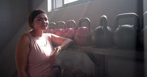 Woman leaning on kettlebell rack checking smartwatch after strength training in sunlit gym