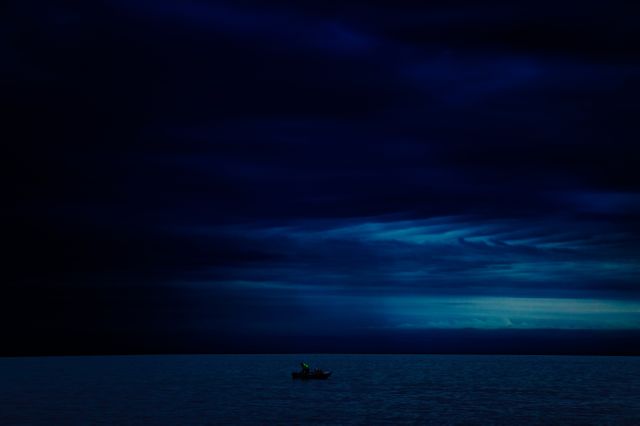 Solitary boat floating on calm sea under a dark, cloudy night sky creates a serene and quiet atmosphere. Perfect for concepts of tranquility, solitude, and reflection. Can be used for calming backgrounds, posters promoting peace or mental wellness, or in articles dealing with the concept of solitude and nature's beauty.