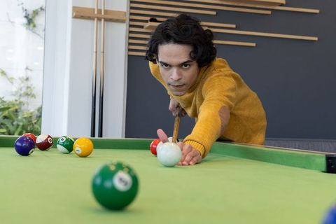 Concentrated young man lining up pool cue on green table in lounge wearing mustard sweater