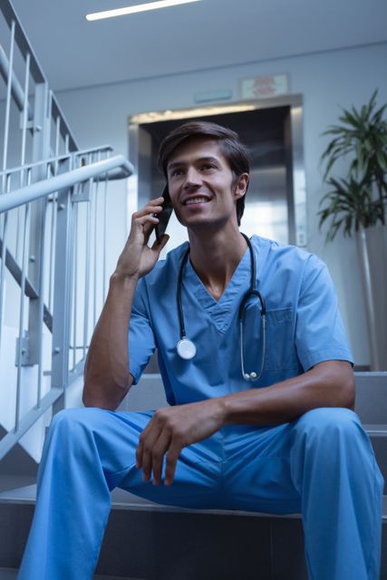 Front view of Caucasian male surgeon talking on mobile phone while sitting on stairs at hospital