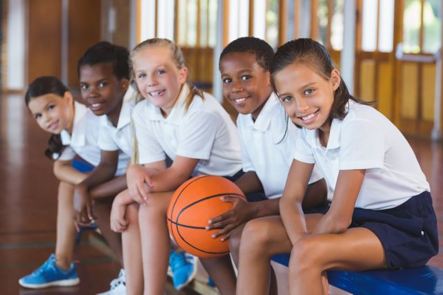 Diverse group of school kids sitting in a basketball court, smiling and holding a basketball. Ideal for educational materials, sports programs, youth activities, and promoting teamwork and diversity in schools.