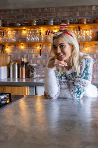 Smiling woman leaning on rustic kitchen counter wearing fuzzy festive sweater and candy-cane headban