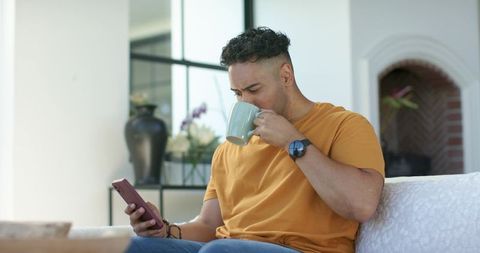 African american man sipping coffee checking smartphone on couch in sunlit living room