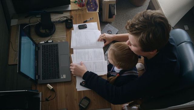 Parent tutoring child at home desk with laptop and workbook for focused study