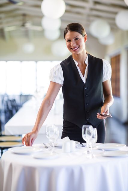 Waitress in uniform setting table in a modern restaurant, smiling warmly. Ideal for use in hospitality industry promotions, restaurant advertisements, service training materials, and customer service brochures.
