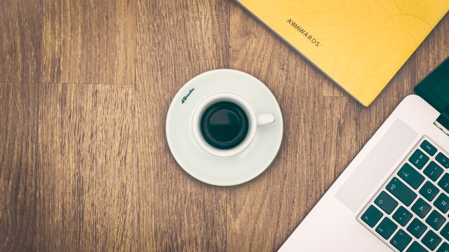 This image depicts a minimalist workspace with a cup of coffee, a laptop, and a notebook on a wooden desk. Ideal for blogs or websites about productivity, remote work, modern office setups, coffee culture, and workspace organization.